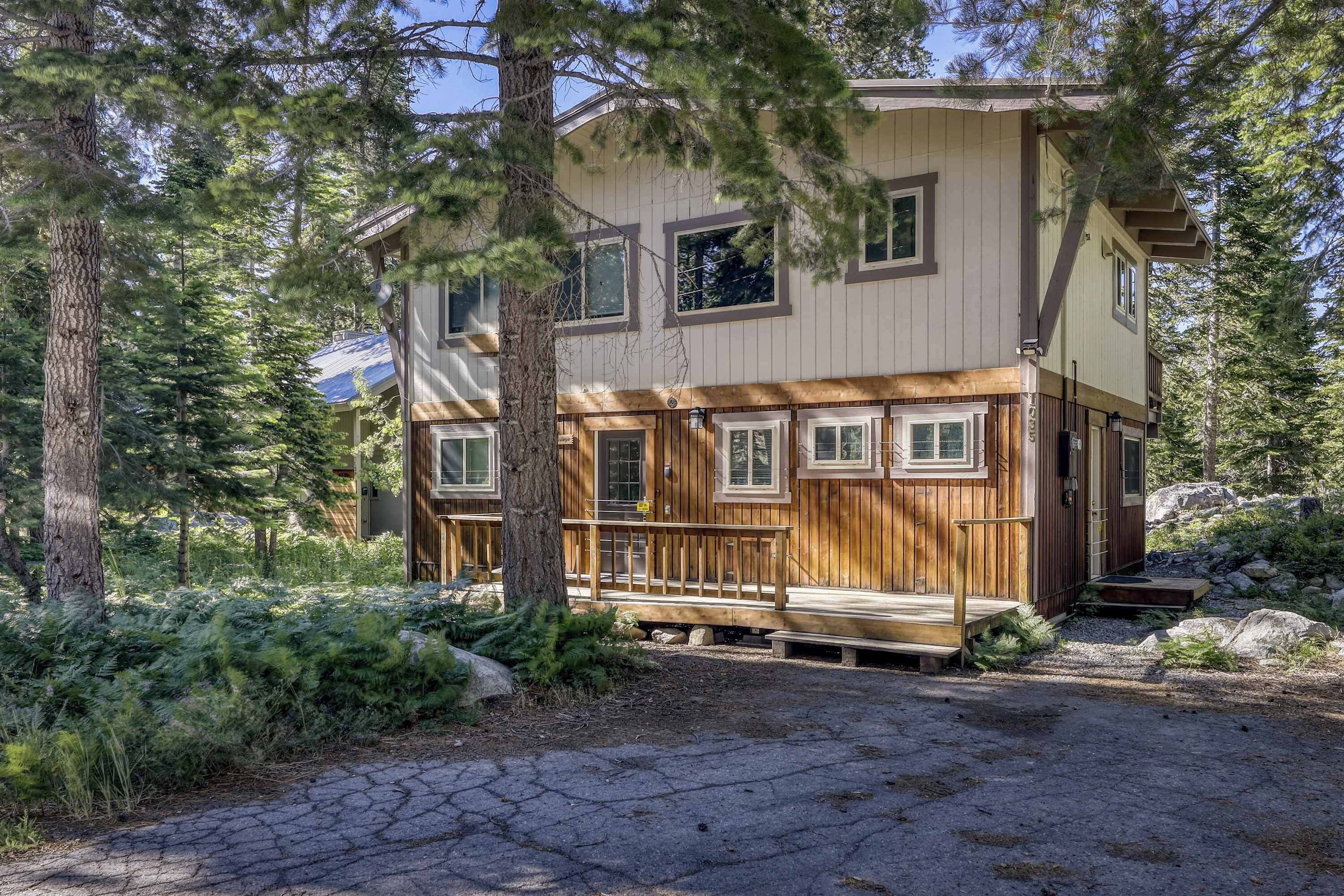 A two-story wooden cabin with a deck, surrounded by trees and a gravel driveway.