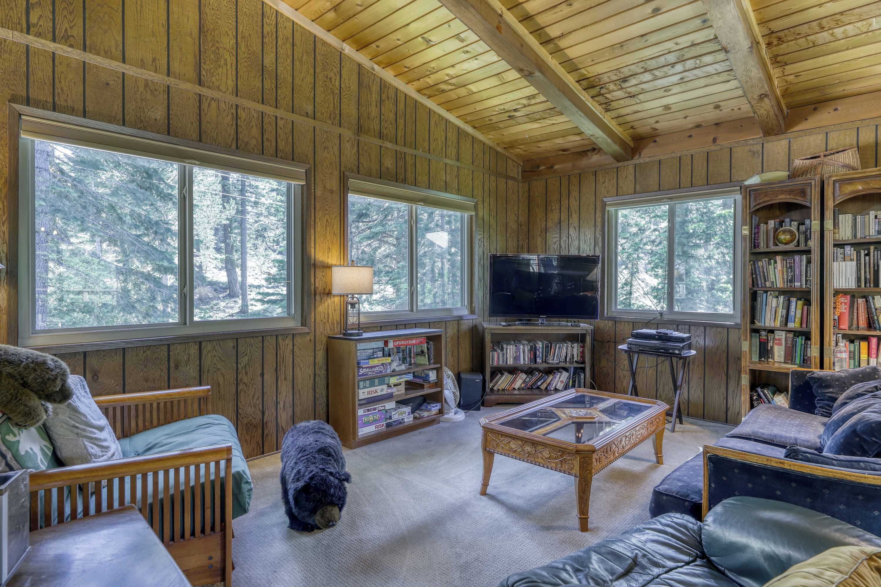 A cozy cabin interior with wooden walls, large windows, a television, a bookshelf, and a dog.