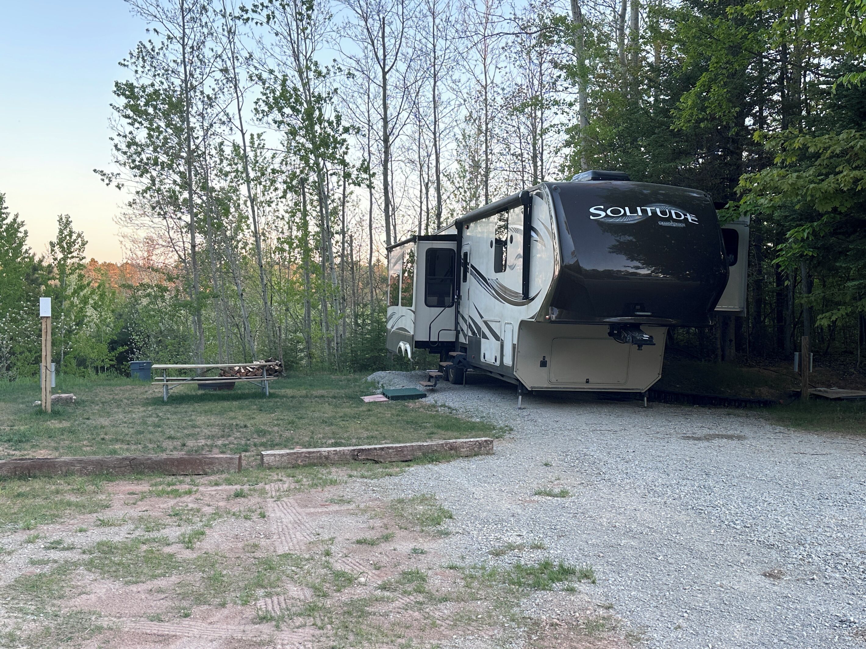A recreational vehicle (RV) parked in a wooded area with a gravel path, surrounded by trees and a picnic table.