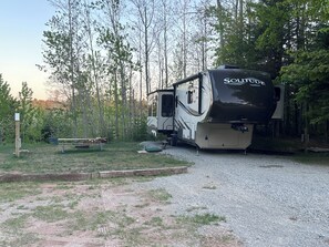 A recreational vehicle (RV) parked in a wooded area with a gravel path, surrounded by trees and a picnic table.