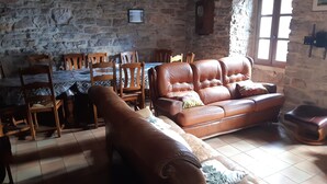 Ein Zimmer mit Steinwänden, einem Ledersofa, Holzstühlen, einem Esstisch und einem Fenster.
