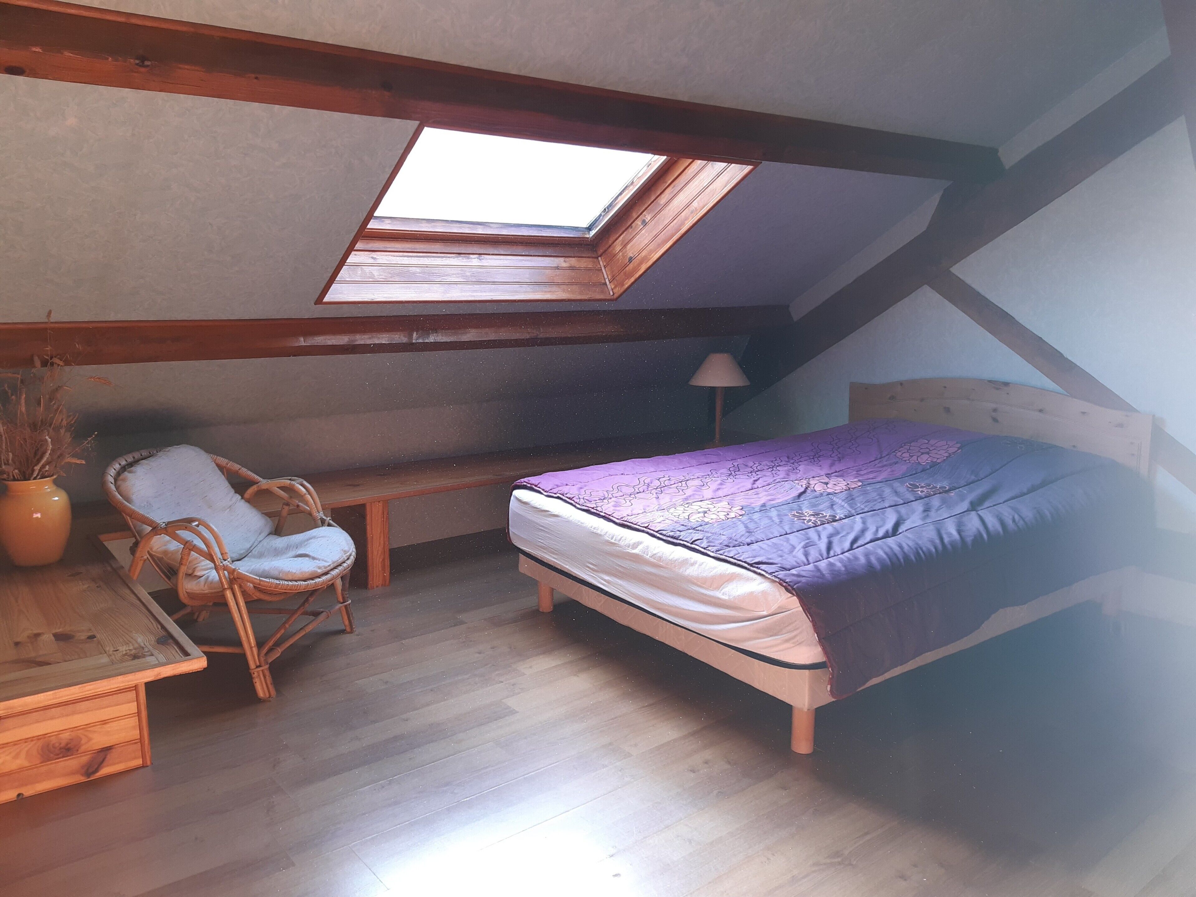 Une chambre à coucher aménagée dans un espace en hauteur, avec une lucarne, un lit, une chaise et un bureau.