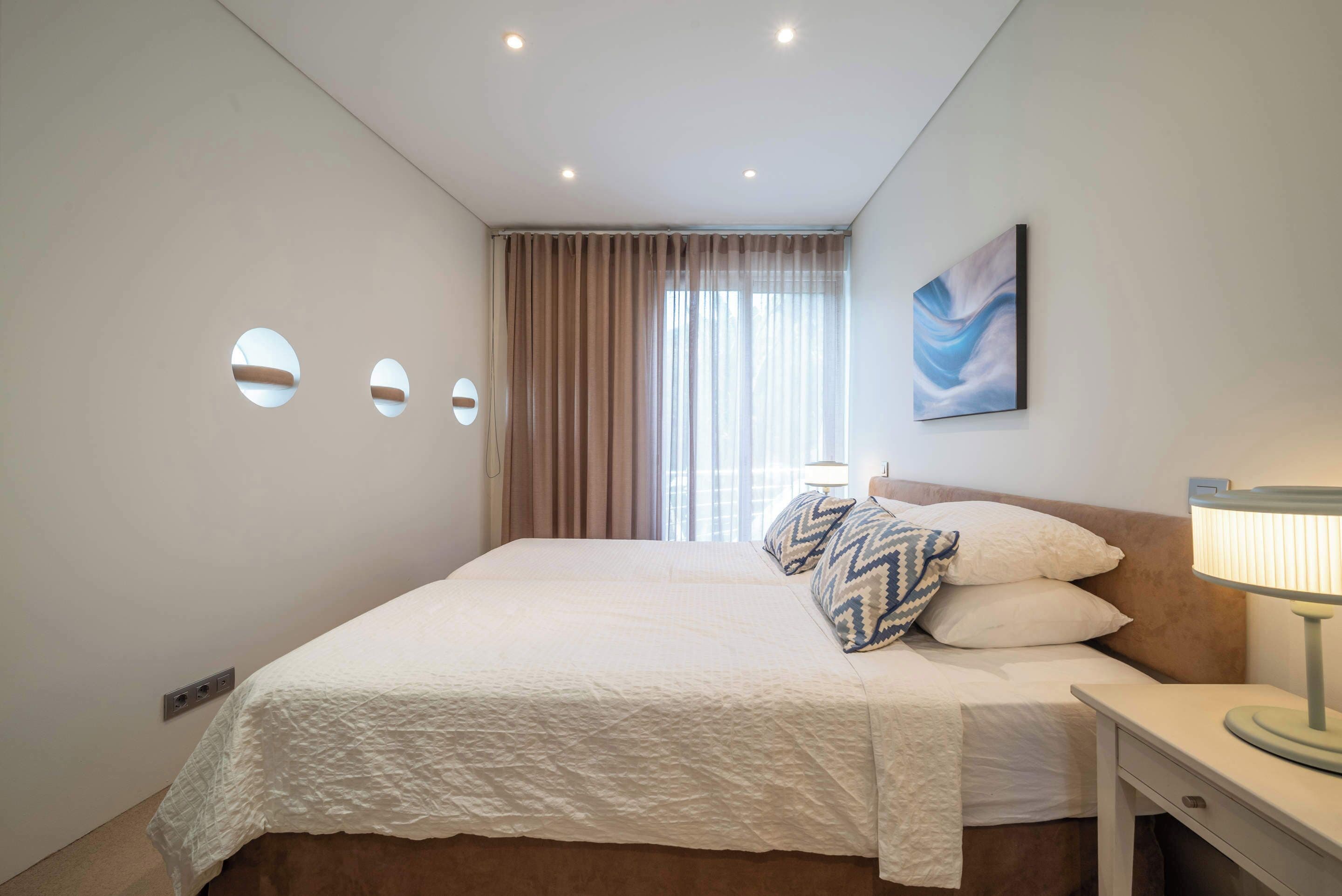 Habitación de hotel con cama, mesita de noche, lámpara, un cuadro y ventana con cortinas.