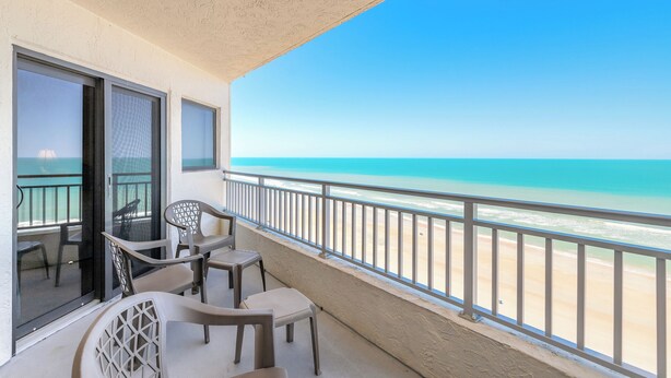 A balcony with a view of the beach, a table with chairs, and a glass door leading to the interior. - 🐟😍Direct Ocean Front, Poolside Play, Panoramic Scene - Seascape Towers 1123🌴 (New Smyrna Beach)