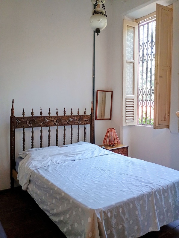 A bedroom with a wooden bed, a bedside lamp, a window with bars, and a mirror on the wall.