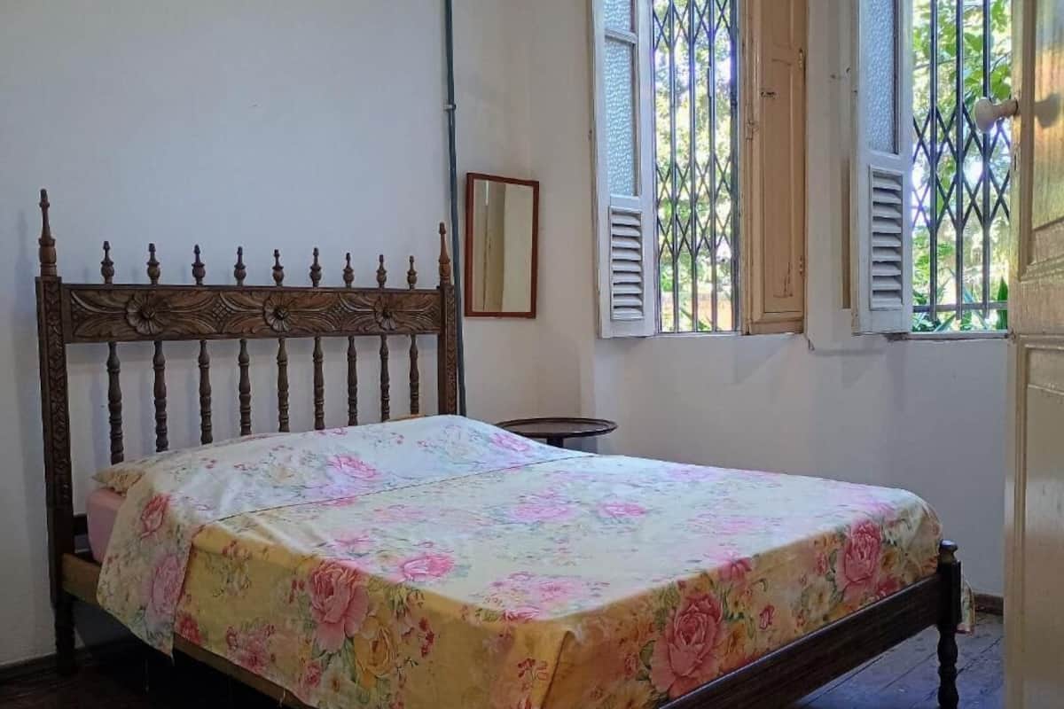 Una cama con colcha floral, un cabecero de madera, una mesita de noche pequeña y una ventana con contraventanas.