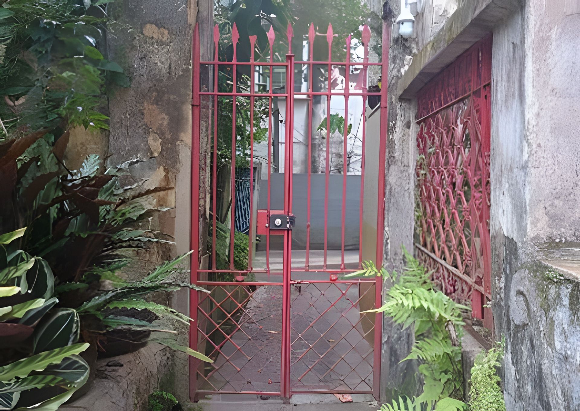 Um portão de metal vermelho com fechadura, dando acesso a um caminho estreito ladeado por vegetação e um edifício com janela vermelha.
