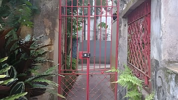 Um portão de metal vermelho com fechadura, dando acesso a um caminho estreito ladeado por vegetação e um edifício com janela vermelha.