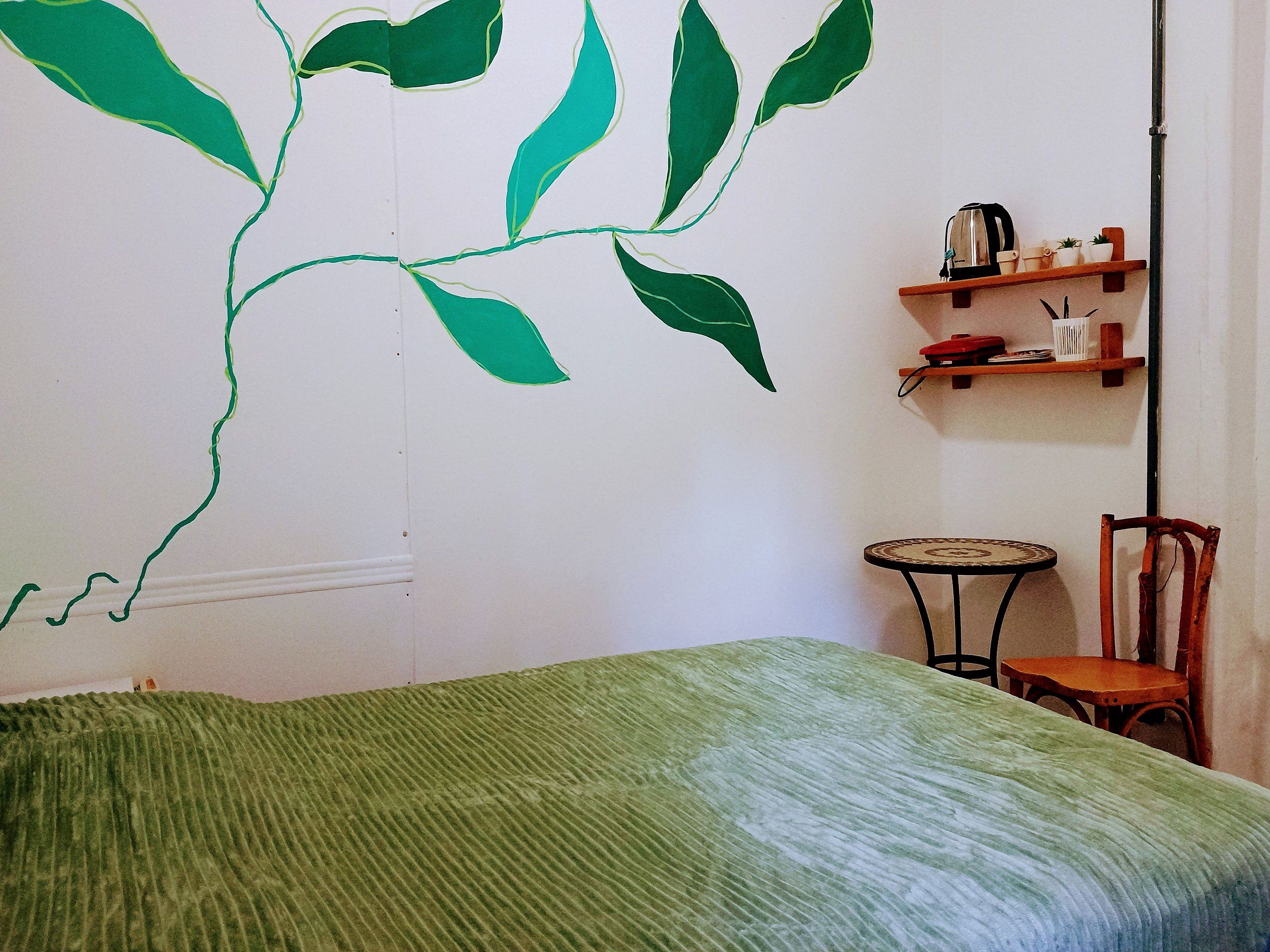 Quarto com cama, cadeira, mesa pequena e parede com padrão de folha verde.