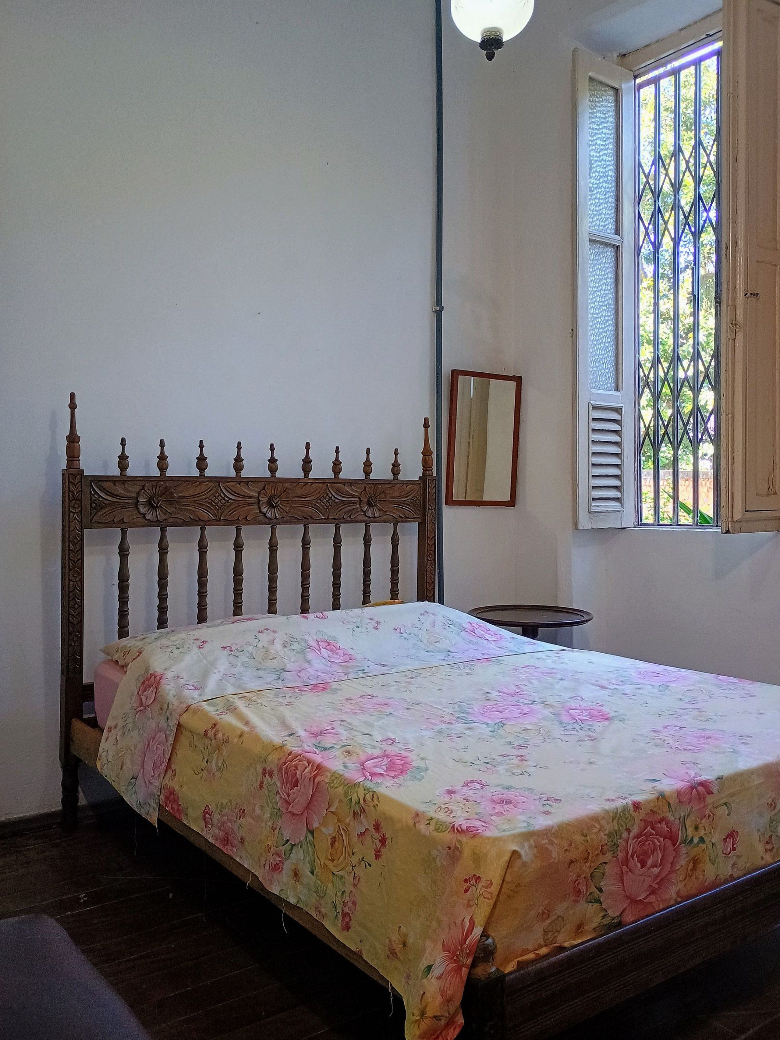 A bedroom with a wooden bed, a bedside table, a mirror, and a window with a view of greenery.