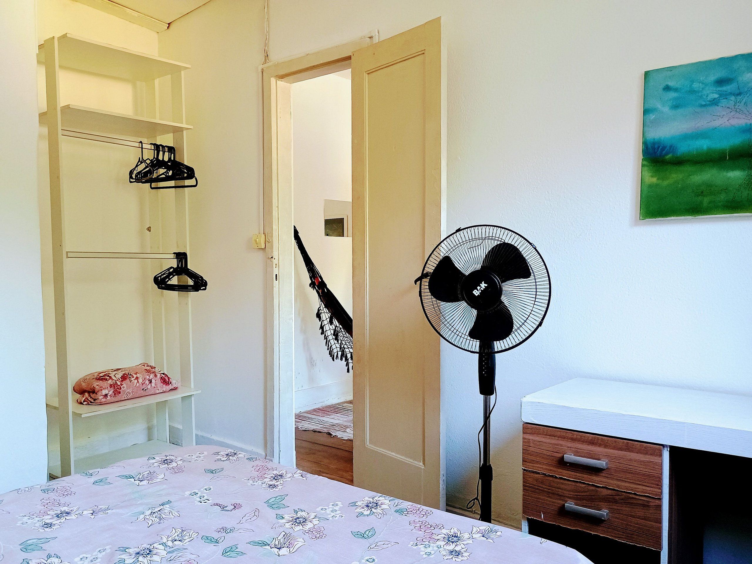 Quarto com cama, ventilador, cômoda de madeira e armário com prateleiras.