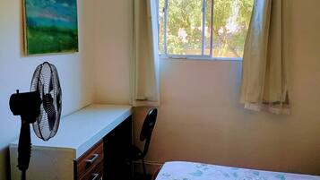A room with a desk, a chair, a standing fan, a window with curtains, and a painting on the wall.