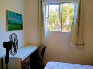 Una habitación con escritorio, silla, ventilador de pie, ventana con cortinas y un cuadro en la pared.