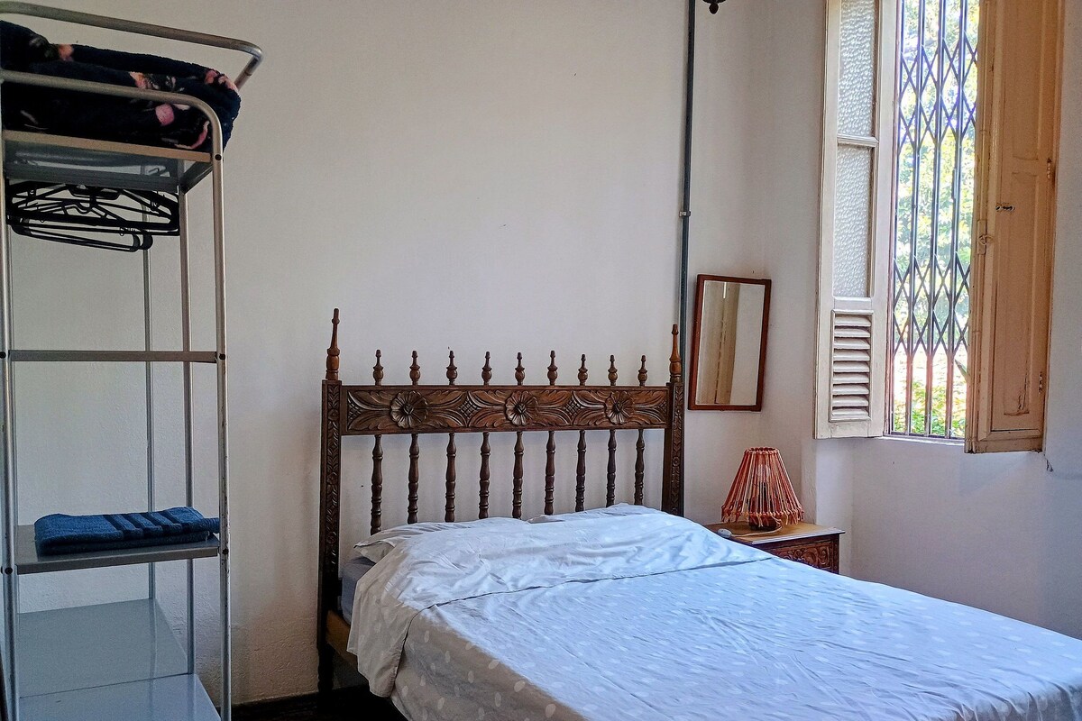 Uma cama de solteiro com cabeceira de madeira, mesa de cabeceira com abajur, janela com grades e estante de metal com roupas.