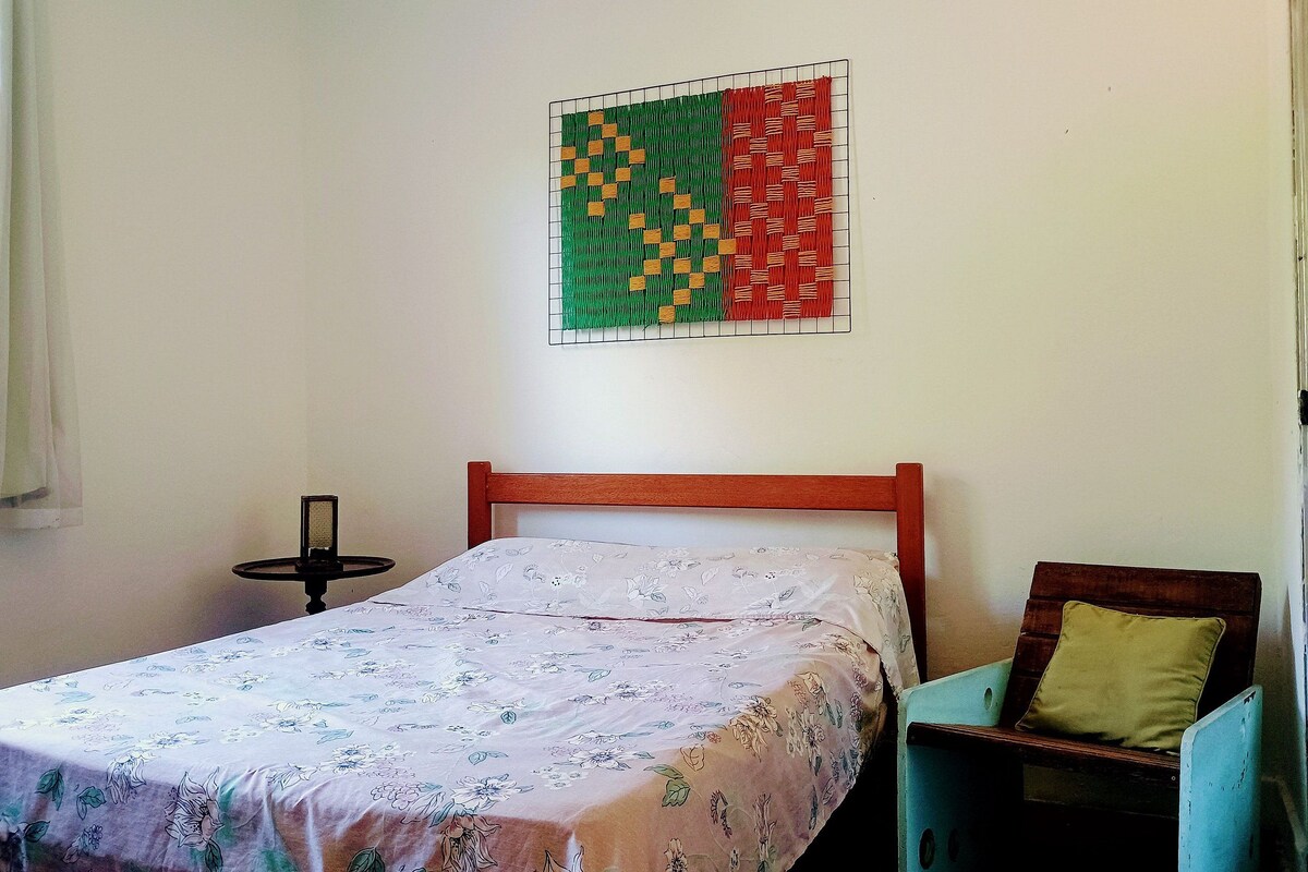 Uma cama de solteiro com colcha florida, cabeceira de madeira, mesa de cabeceira pequena e uma obra de arte pendurada na parede.