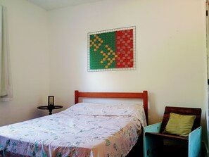 A single bed with a floral bedspread, a wooden headboard, a small bedside table, and a wall-mounted artwork. - Quintal Oceano (Rio de Janeiro)
