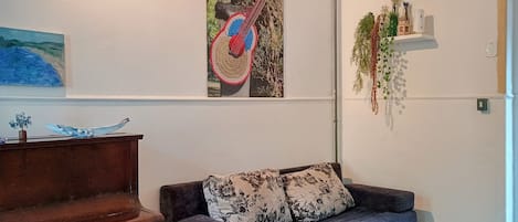 A room with a piano, a sofa with floral cushions, and wall art including a guitar and a painting.