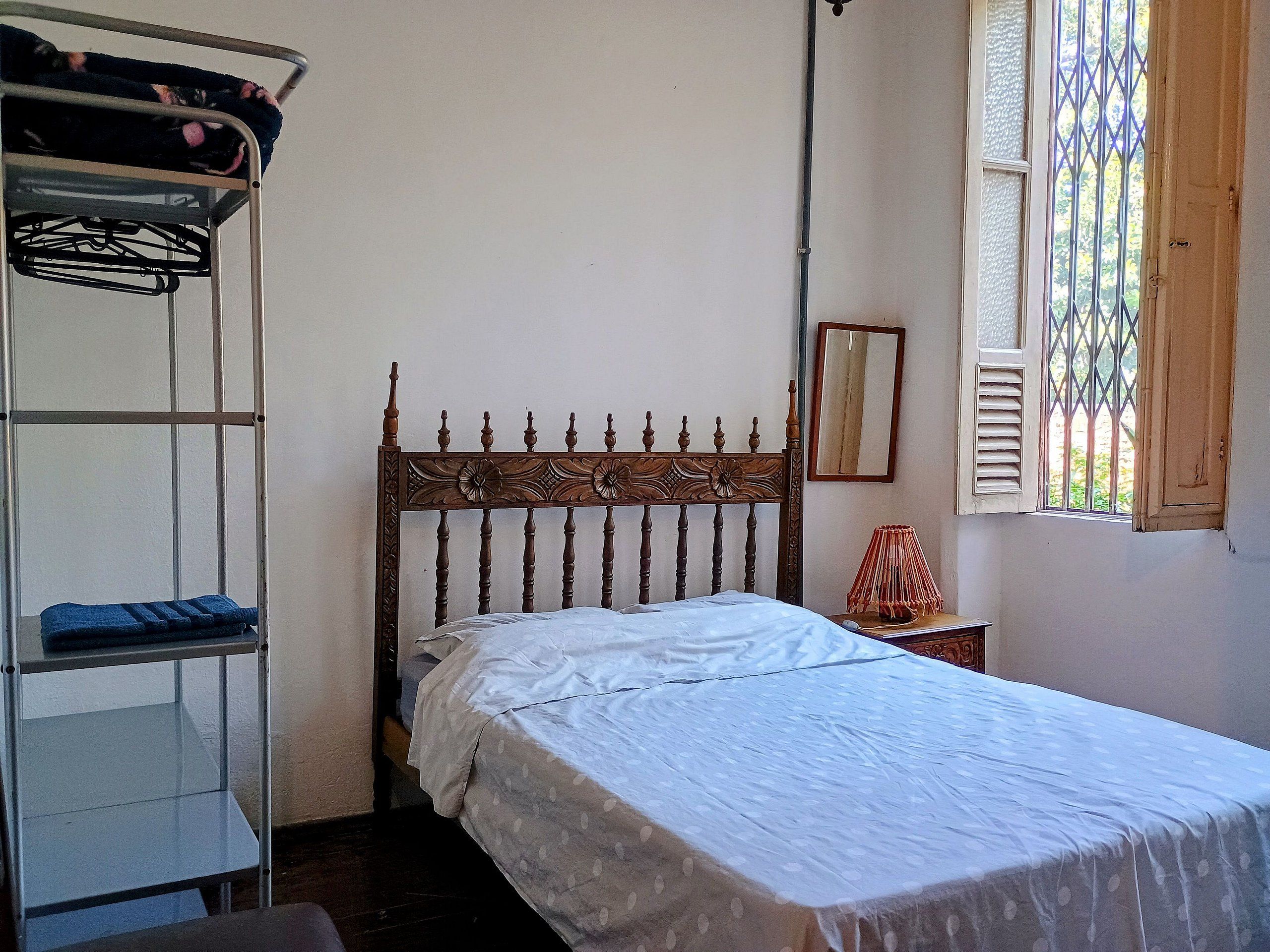 Una cama individual con cabecera de madera, una mesita de noche con lámpara, una ventana con rejas y un estante metálico con ropa.