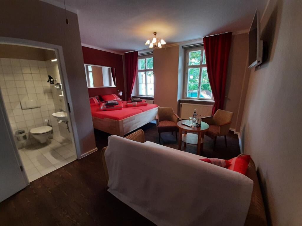 Ein Hotelzimmer mit einem Bett, zwei Stühlen, einem kleinen Tisch, einem Fenster mit Vorhängen und einem durchgegangenen Tür sichtbar liegenden Badezimmer.