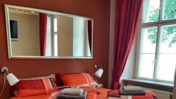 A double bed with a wooden frame and orange bedding, a large mirror above it, and a window with red curtains.