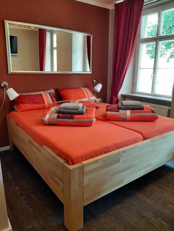 A double bed with a wooden frame and orange bedding, a large mirror above it, and a window with red curtains.