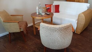 A living room with a white sofa, two patterned armchairs, a small round table with a bottle and glass, and a wooden cabinet in the background.
