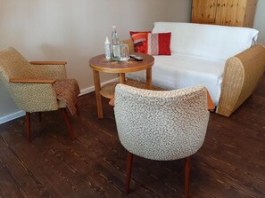 A living room with a white sofa, two patterned armchairs, a small round table with a bottle and glass, and a wooden cabinet in the background.