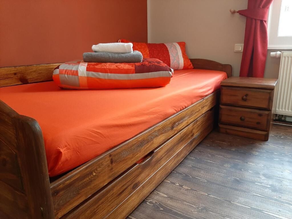 A wooden bed with an orange bedspread, a wooden nightstand with a drawer, and a red curtain.