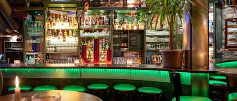 Un espace bar avec des tabourets, des étagères remplies de bouteilles et un comptoir illuminé de lumière verte.