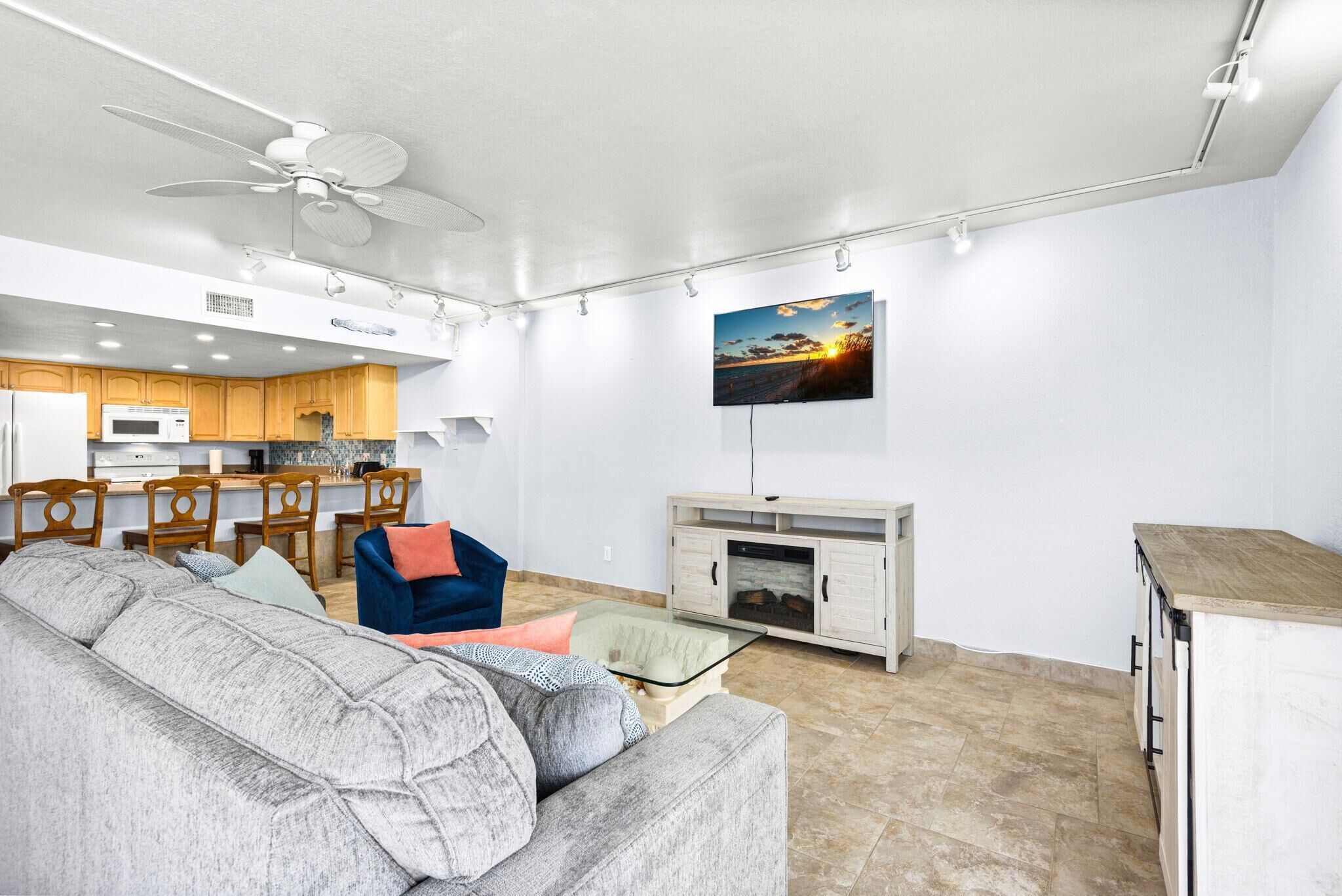 Uma sala de estar moderna com um sofá, uma mesa de centro de vidro, uma TV fixada na parede e uma área de cozinha com armários de madeira e um micro-ondas.