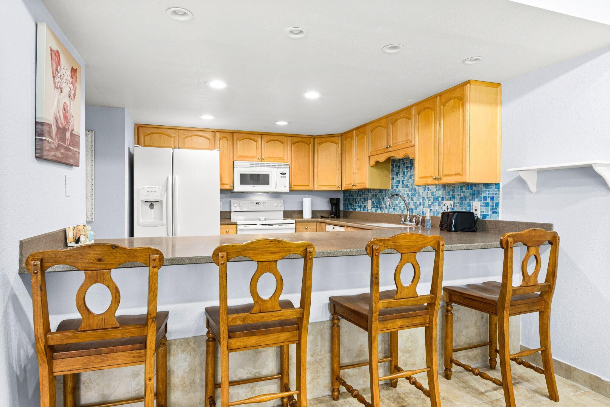 A kitchen with wooden cabinets, a white refrigerator, a microwave, a stove, and a tiled backsplash. There are three wooden chairs at a kitchen island.