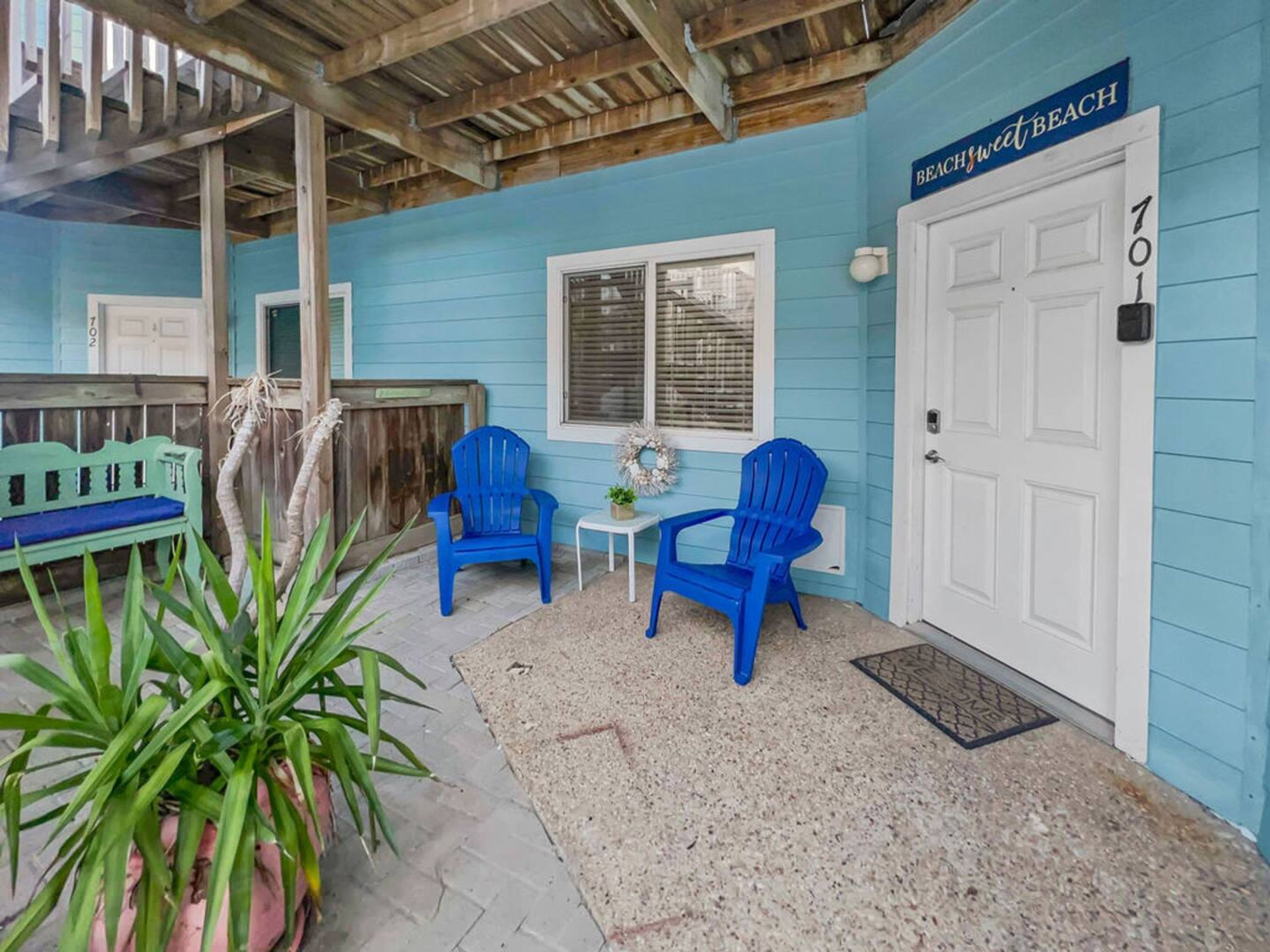 A covered porch with a blue chair, a small table, and a door labeled '701 BEACHSWEET BEACH.'