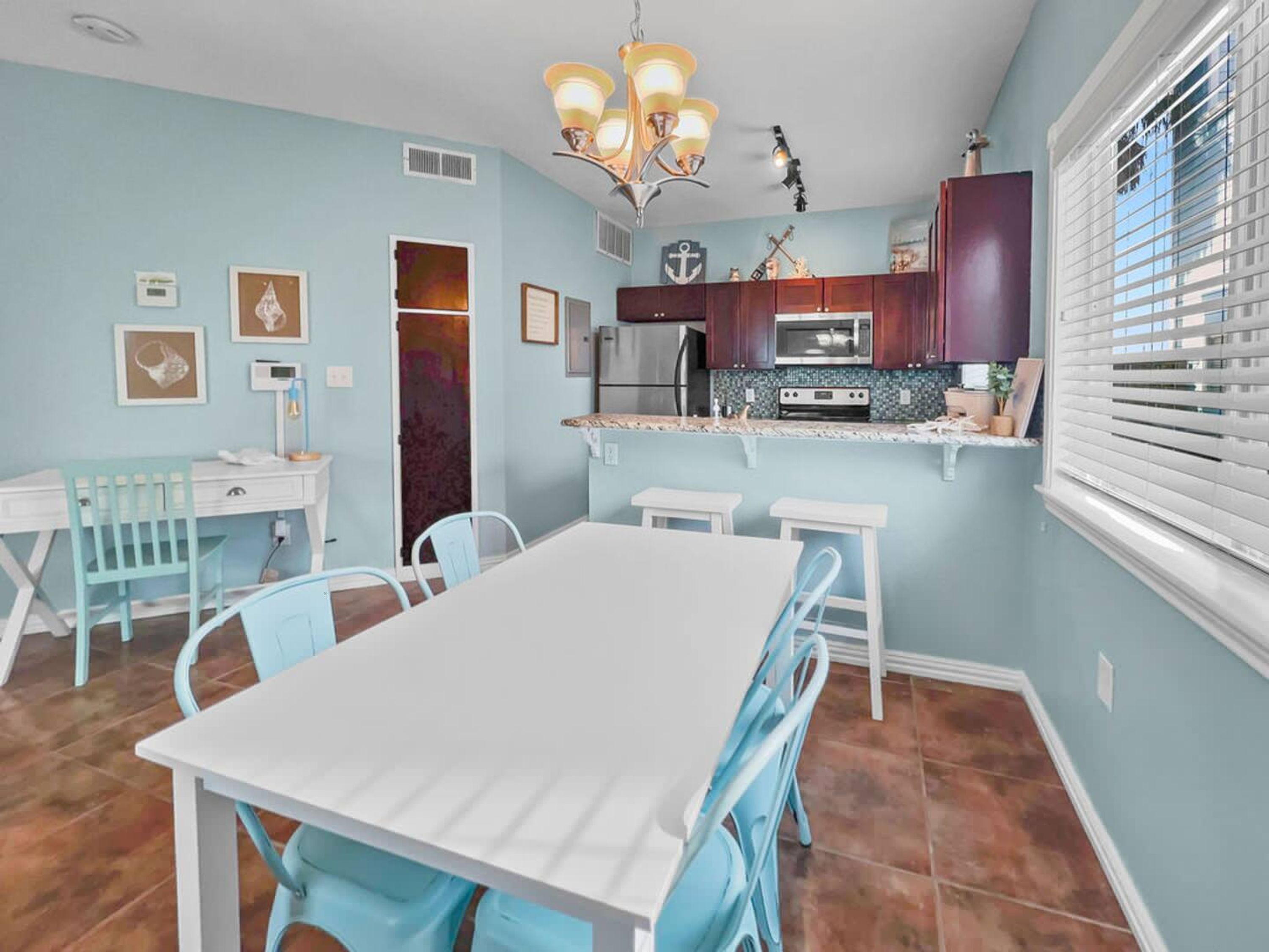 Een eetgedeelte met een witte tafel en blauwe stoelen, een keuken met donkere kasten en een magnetron, en een raam met zonwering.
