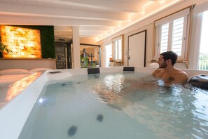 A man in a hot tub in a modern room with a large window, a wall-mounted heater, and a visible kitchen area. - Just Relax Home (Sant'Angelo Romano)