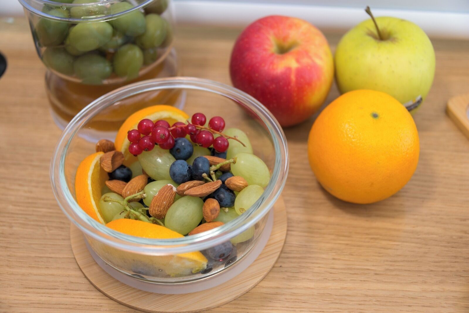 Un cuenco de vidrio transparente con frutas y nueces mezcladas sobre una superficie de madera, acompañado de frutas enteras y un tarro con uvas al fondo.