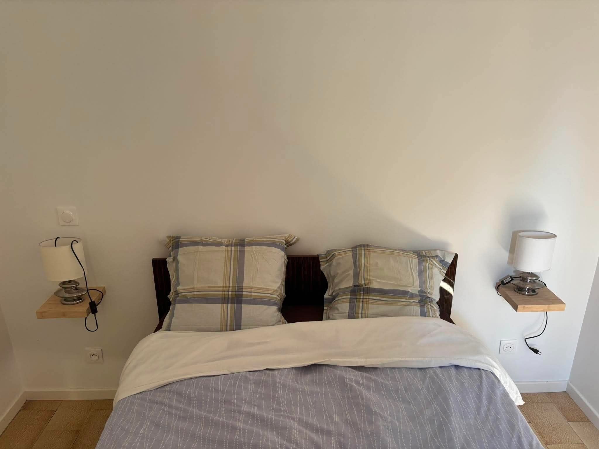 A bedroom with a bed, two pillows, a bedside table with a lamp, and a power outlet.