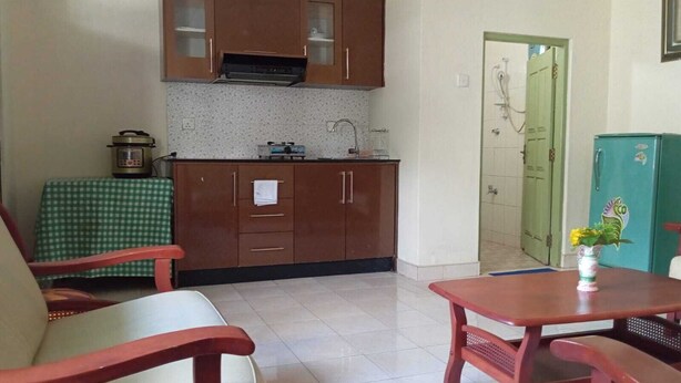 Uma cozinha compacta com fogão, pia e armários, uma mesa de jantar com bancos, e uma porta verde que dá para um banheiro.