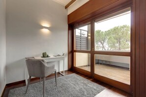 Ein Zimmer mit Schreibtisch, Stuhl und großem Fenster mit Blick auf Bäume.