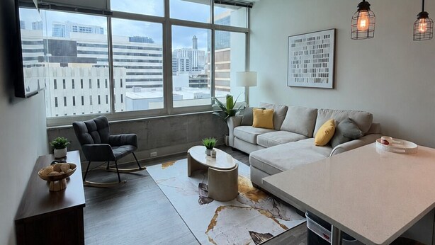 Ein modernes Wohnzimmer mit einer Couch, einem Couchtisch, einem Fernseher und Blick auf die Stadt durch große Fenster.