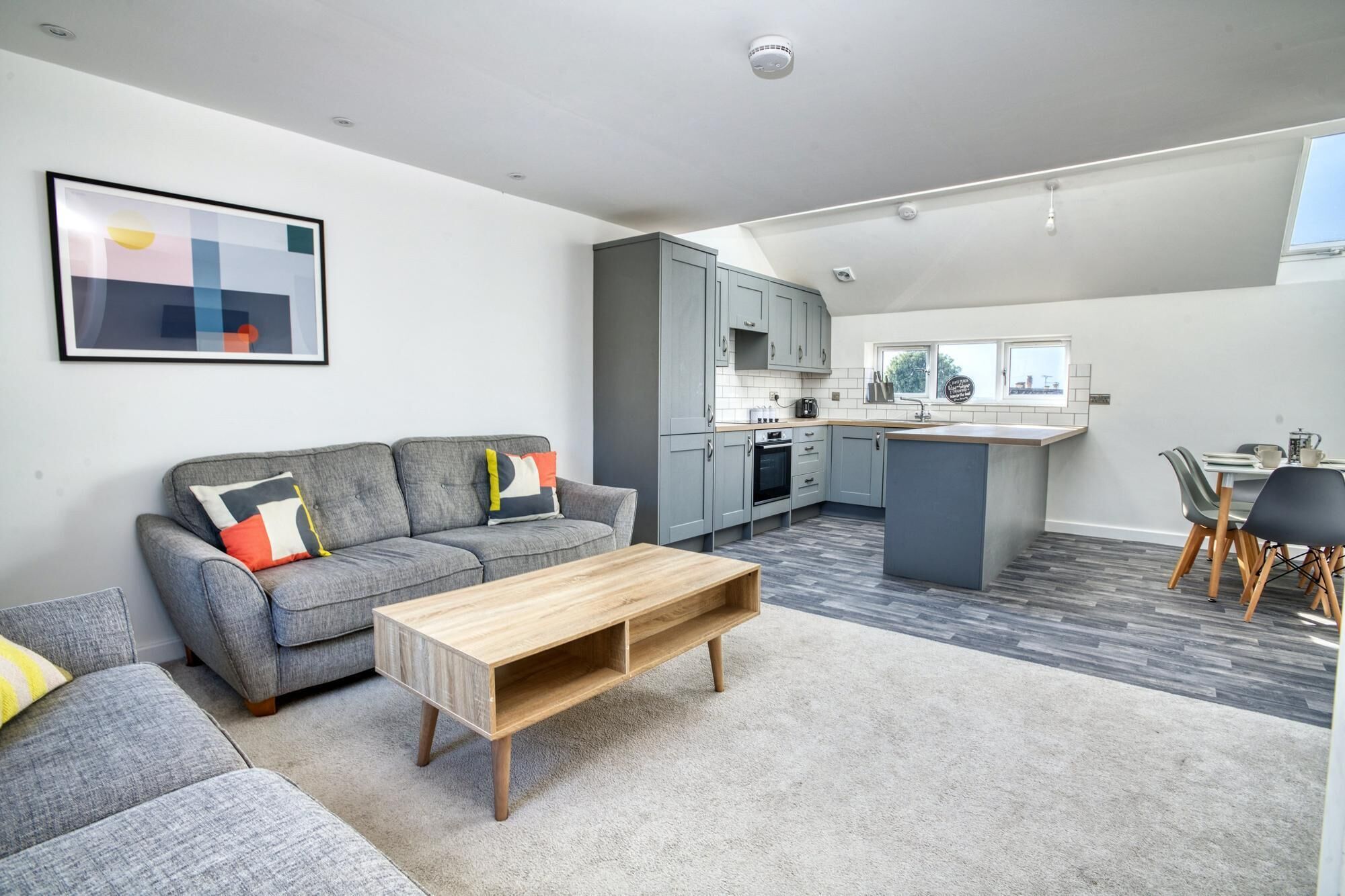 A modern apartment with a grey sofa, a wooden coffee table, a kitchen with grey cabinets, and a dining area with a table and chairs.