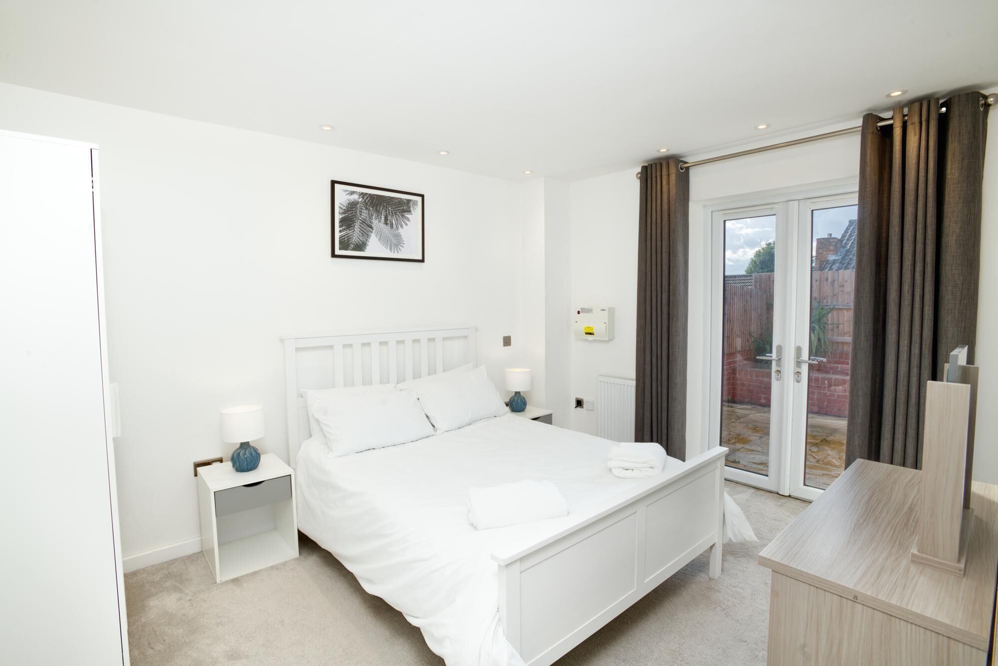 A bedroom with a white bed, bedside tables, a framed picture on the wall, and a sliding glass door leading to an outdoor area.