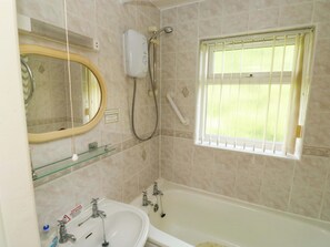A bathroom with a bathtub, a sink, a mirror, a showerhead, and a window with blinds. - Cottage 28 (Brixham)