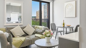 Ein modernes Wohnzimmer mit einer Couch, einem kleinen runden Tisch mit einer Flasche und Gläsern, einer Topfpflanze und einem großen Fenster mit Blick auf die Stadt.