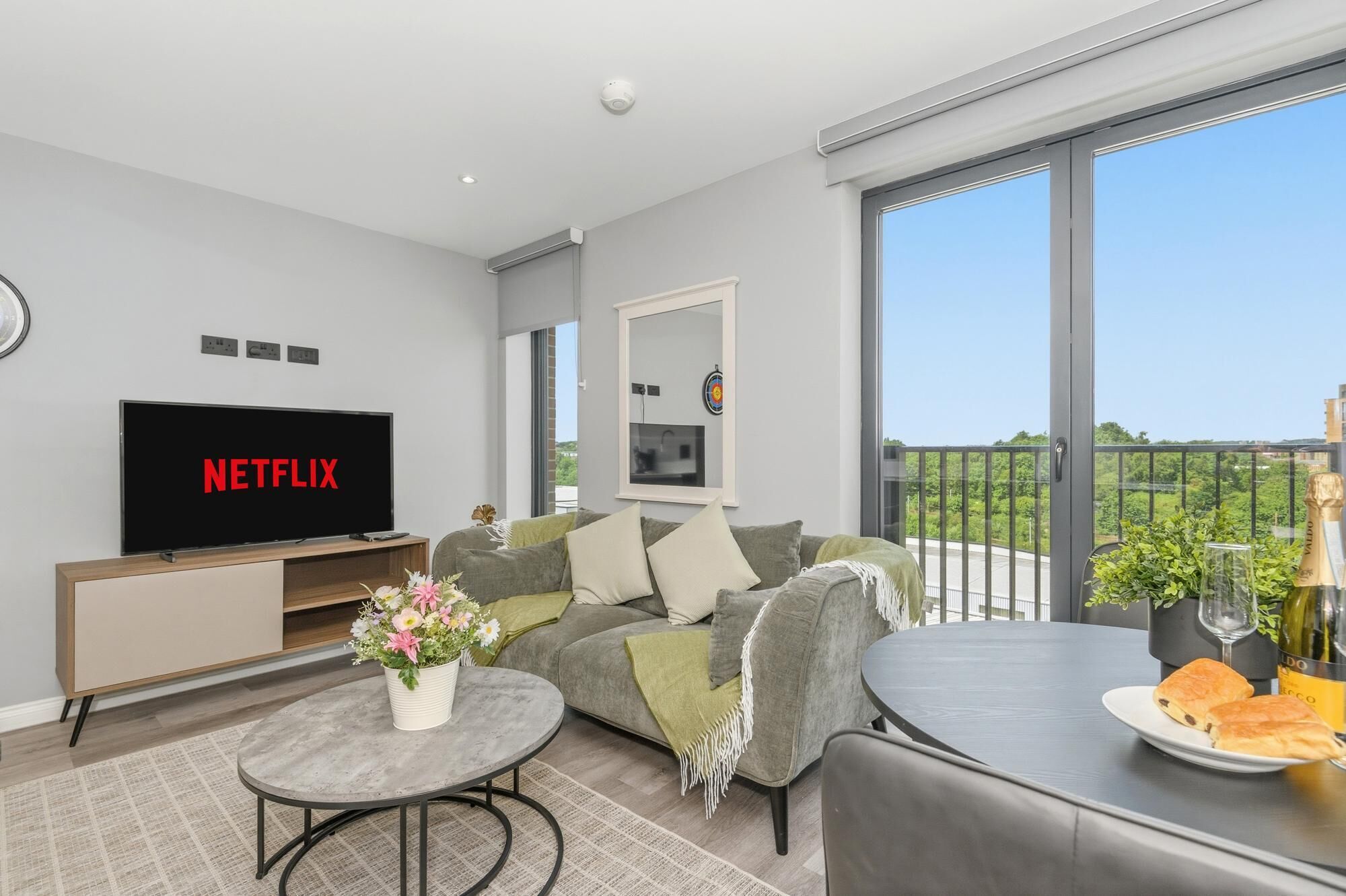 A modern living room with a flat-screen TV, a sofa, a coffee table, and a balcony with a view.