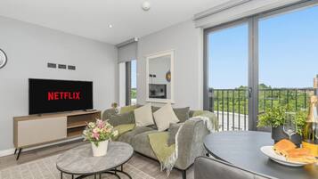 A modern living room with a flat-screen TV, a sofa, a coffee table, and a balcony with a view.