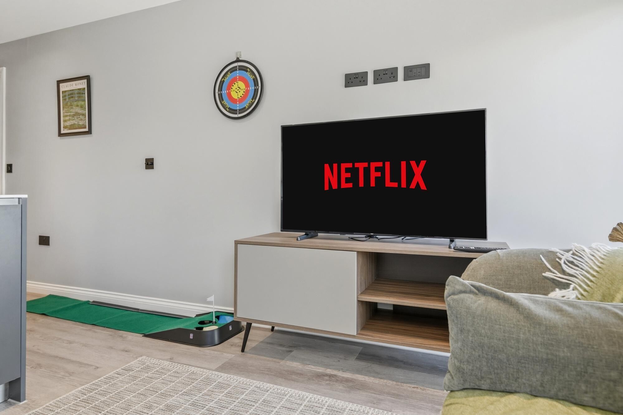 Un saló modern amb televisió plana que mostra 'NETFLIX' en lletres vermelles, un joc de fletxes i una fotografia encadrada a la paret.