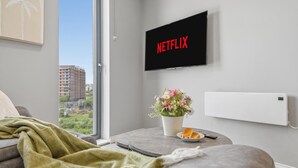Una sala de estar moderna con un televisor mostrando 'NETFLIX', una mesita con un florero, una manta y un plato con pan.