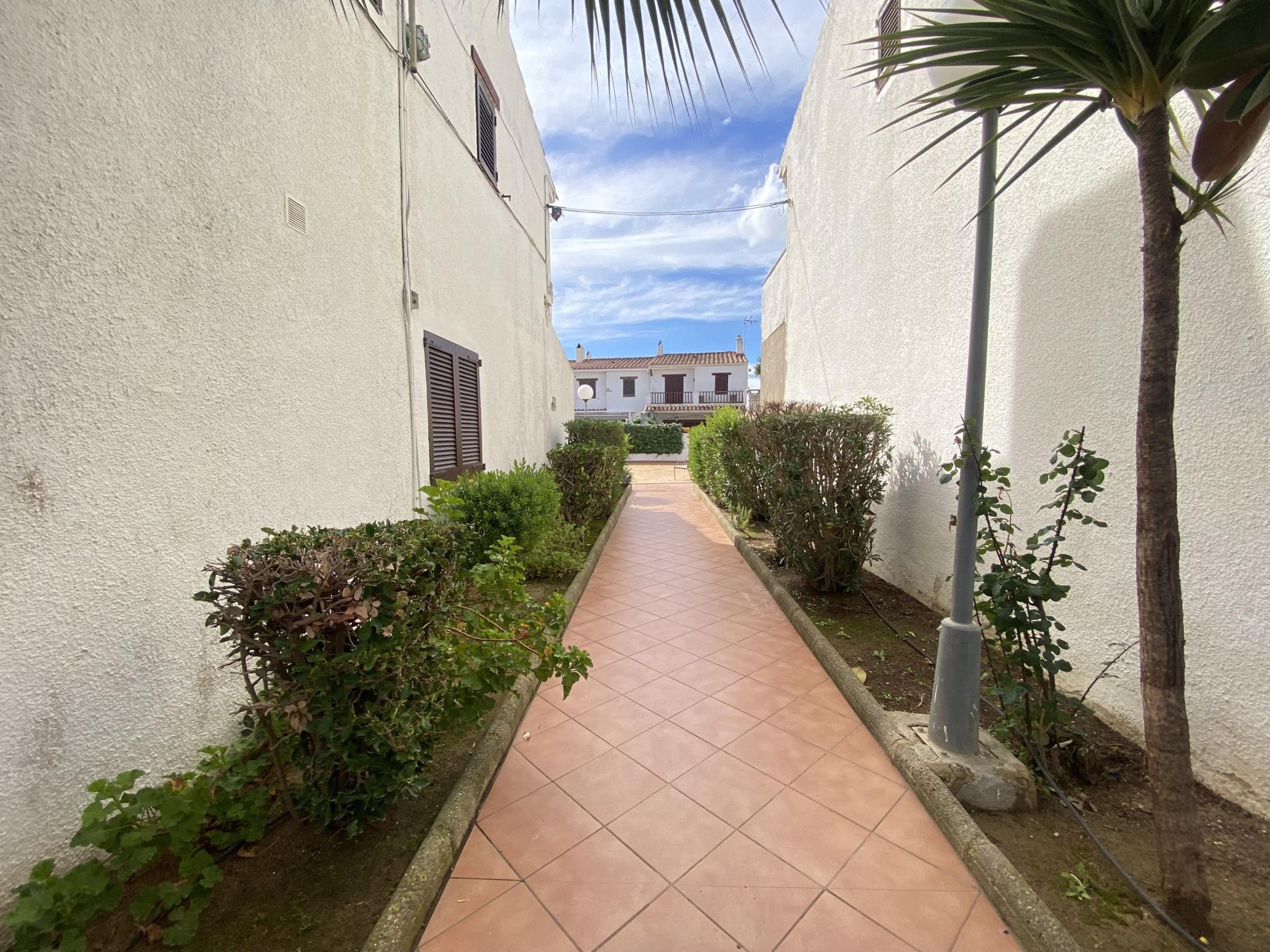 A narrow pathway flanked by white buildings with shutters, leading to a distant house.