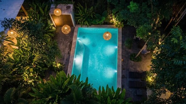 Una piscina rectangular rodeada de abundante vegetación y estructuras de piedra, con luces colgantes que brindan iluminación.
