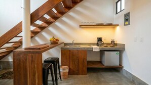 Cocina con lavamanos, cafetera y un refrigerador peque ño. Se observa una escalera de madera y un taburete.